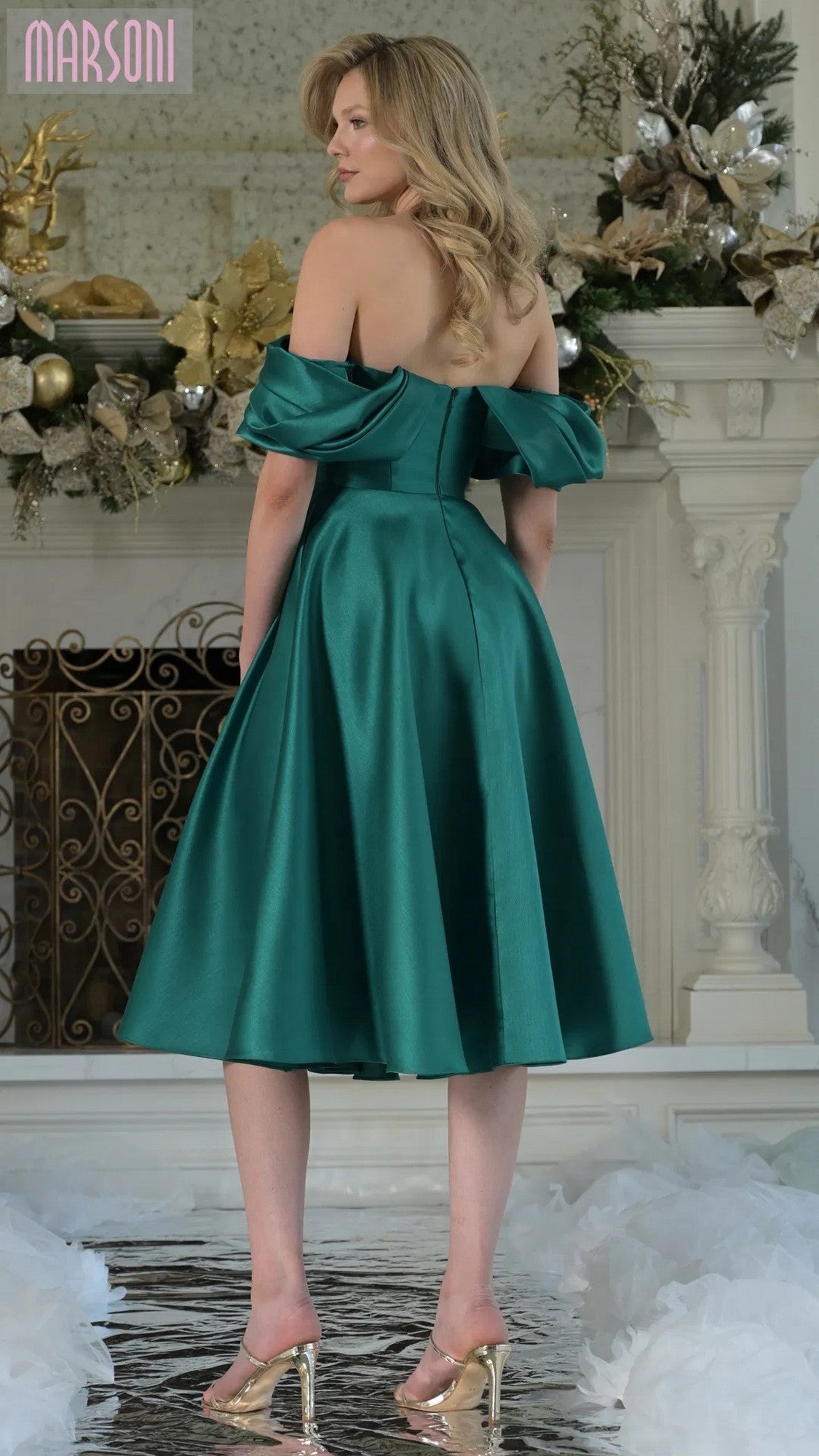 A woman in the Marsoni MV1369 knee-length semi-formal dress with clear heels stands on a reflective floor before a decorated fireplace.